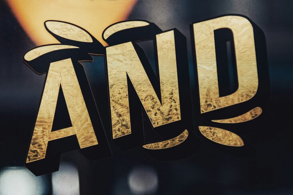 Word "AND" in gold letters, in relief, on a Word "AND" in gold letters on a dark background, representing the transition from a binary "or" logic to an integrative "and" posture.