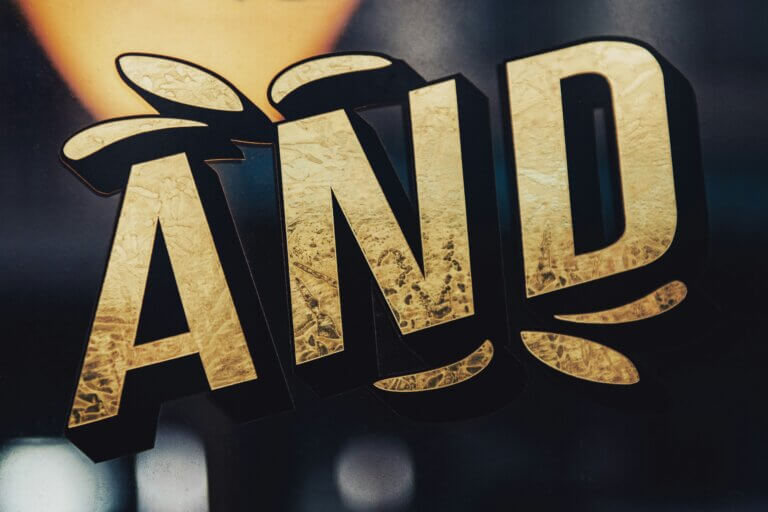 Word "AND" in gold letters, in relief, on a Word "AND" in gold letters on a dark background, representing the transition from a binary "or" logic to an integrative "and" posture.