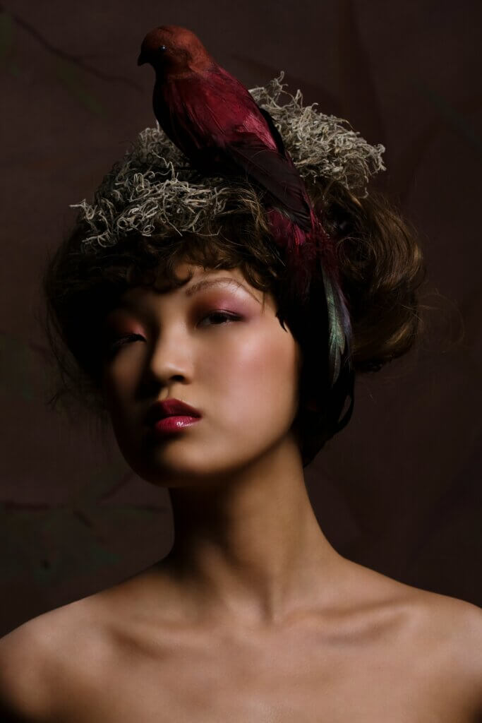 We see a woman of Asian origin, with a direct but inscrutable gaze. Her head carriage is haughty, her presence immediate. Her hairstyle is not a fixed architecture, but an organic ecosystem where natural fibers (it looks like moss) and an iridescent bird feather are intertwined.